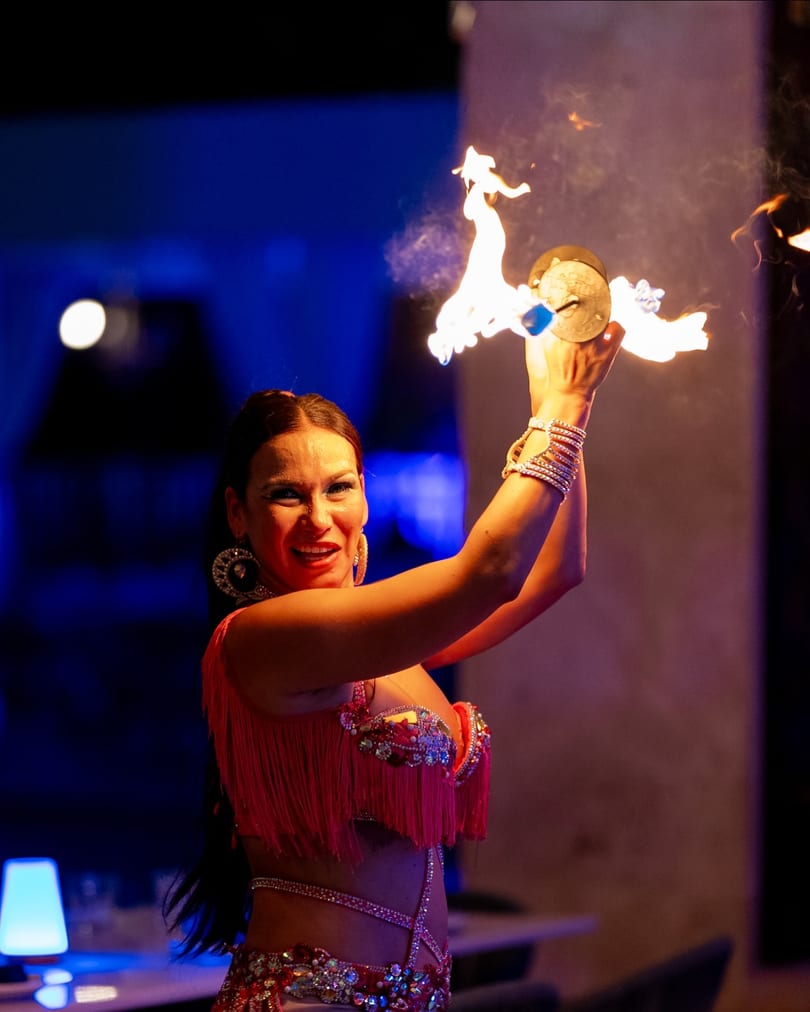 A smiling woman in a vibrant costume performs a fire dance with cymbals, illuminated by blue light.