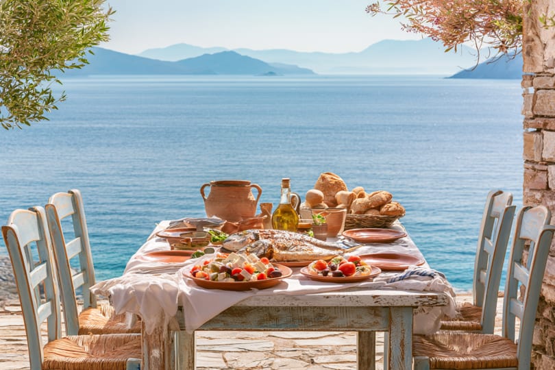 A rustic table set with Mediterranean food, including whole fish and salads, overlooking a blue sea.