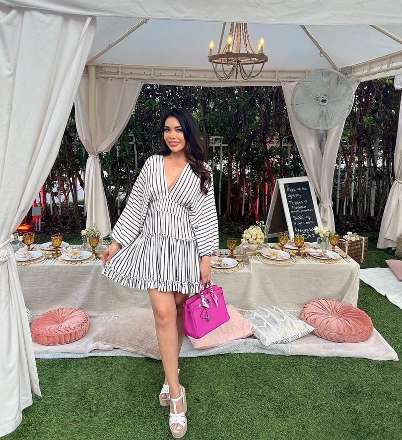 Woman in a striped dress holding a pink purse, standing by a picnic table under a white tent.