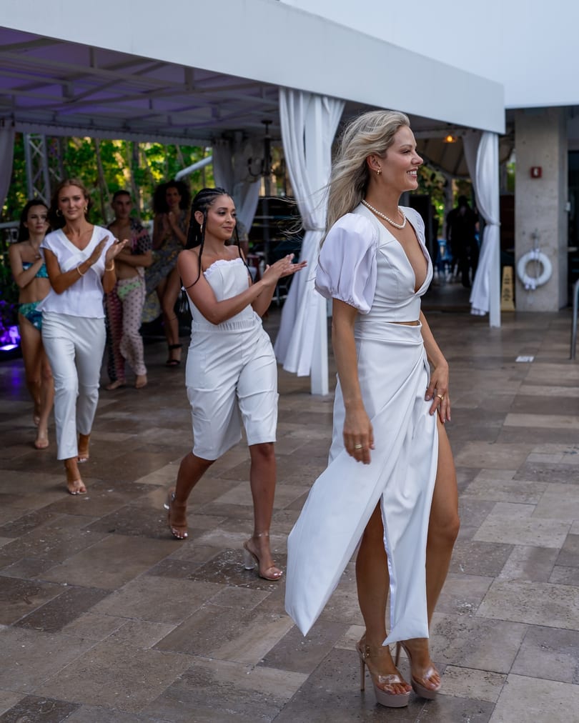 Models in white outfits and clear heels walk on a patio, with others clapping in the background.