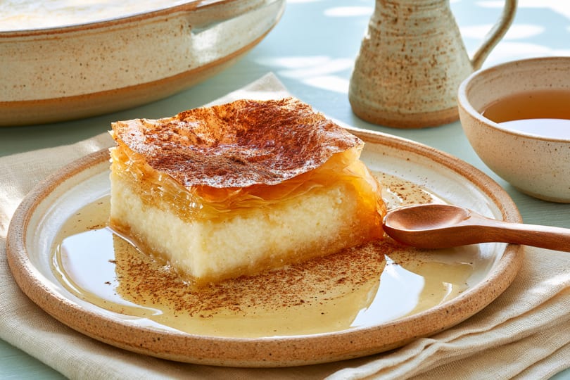 A square piece of bougatsa with honey and cinnamon on a ceramic plate with a wooden spoon.