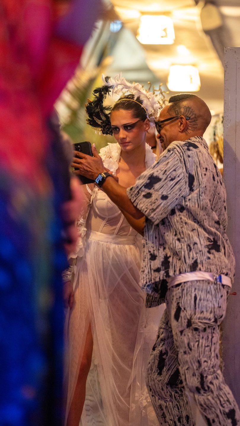 A model in a white feathered headpiece and sheer dress looks at a phone held by a man.