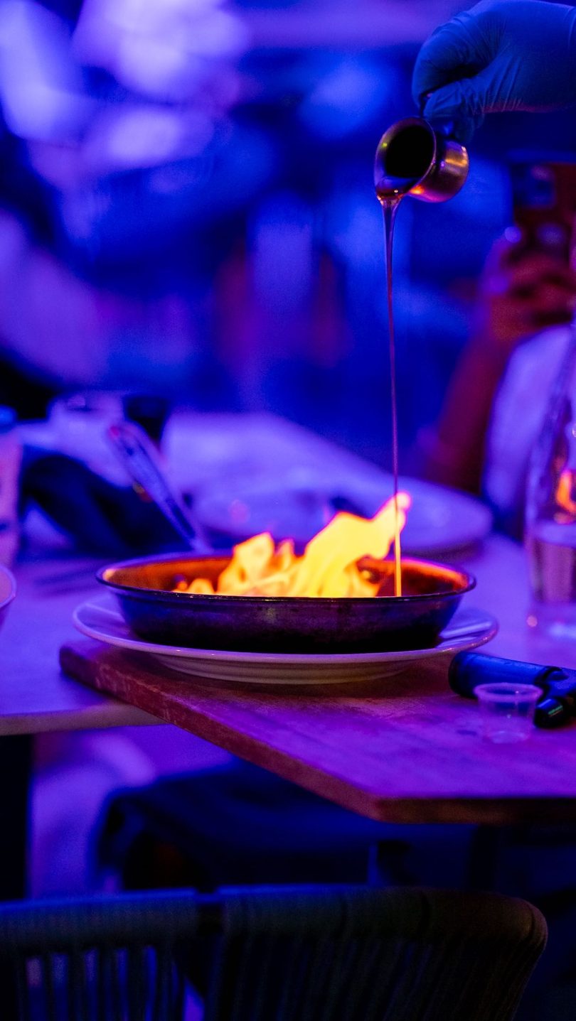 A gloved hand pours liquid from a jigger onto a flaming dish in a dark, blue-lit restaurant.