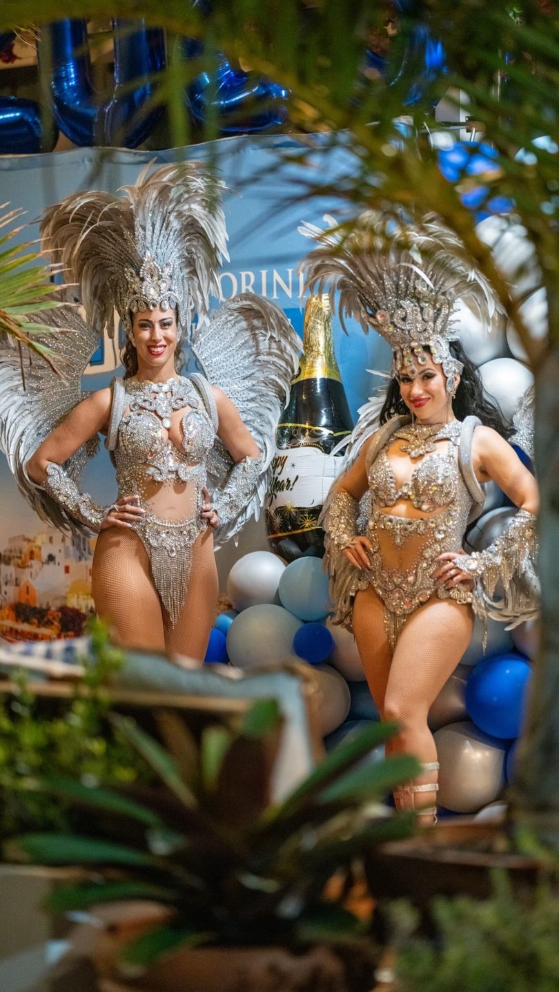 Two women in elaborate silver carnival costumes with feathers and sequins pose at a party.