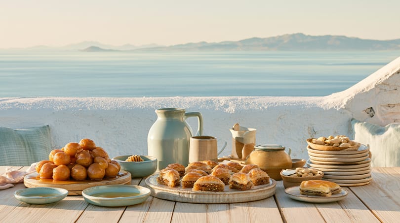 Loukoumades and baklava on a table overlooking the sea and mountains at sunset.