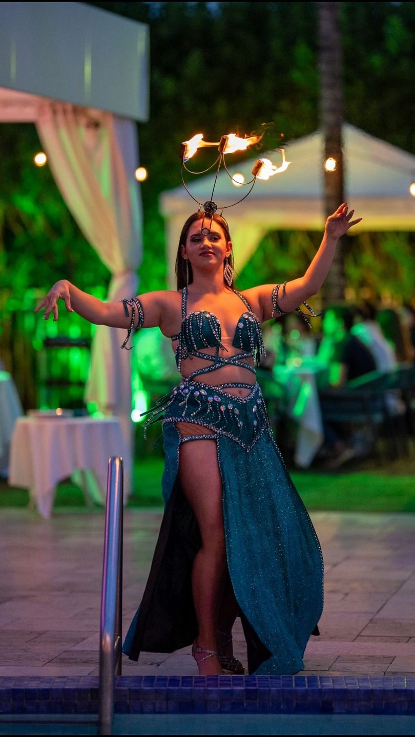 Belly dancer in a teal costume with a flaming candelabra on her head, performing outdoors at night.
